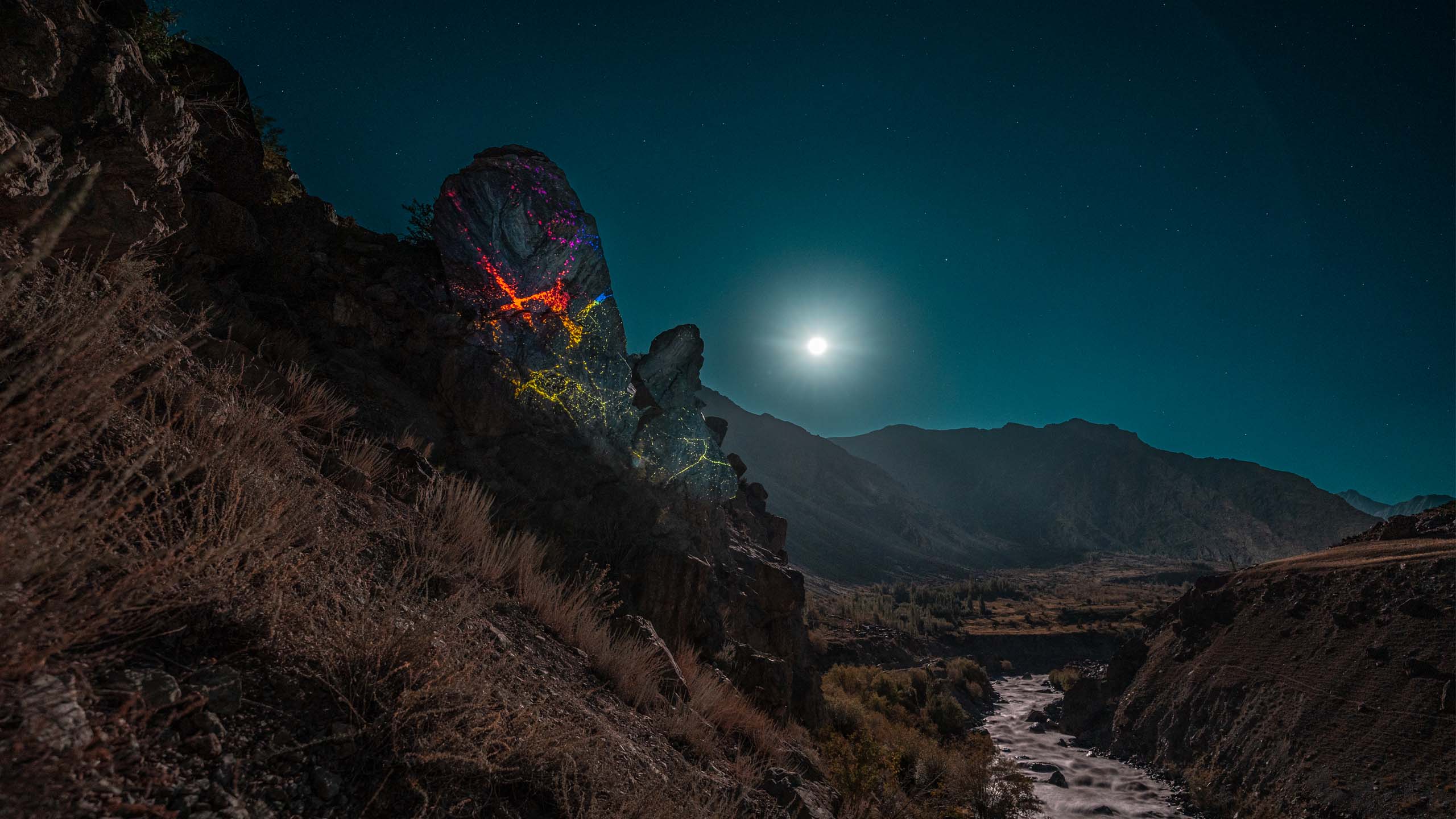 Colorful land-art installation at night on a boulder with stars in the sky and a river and mountains in the baclground. Ladakh, Himalaya