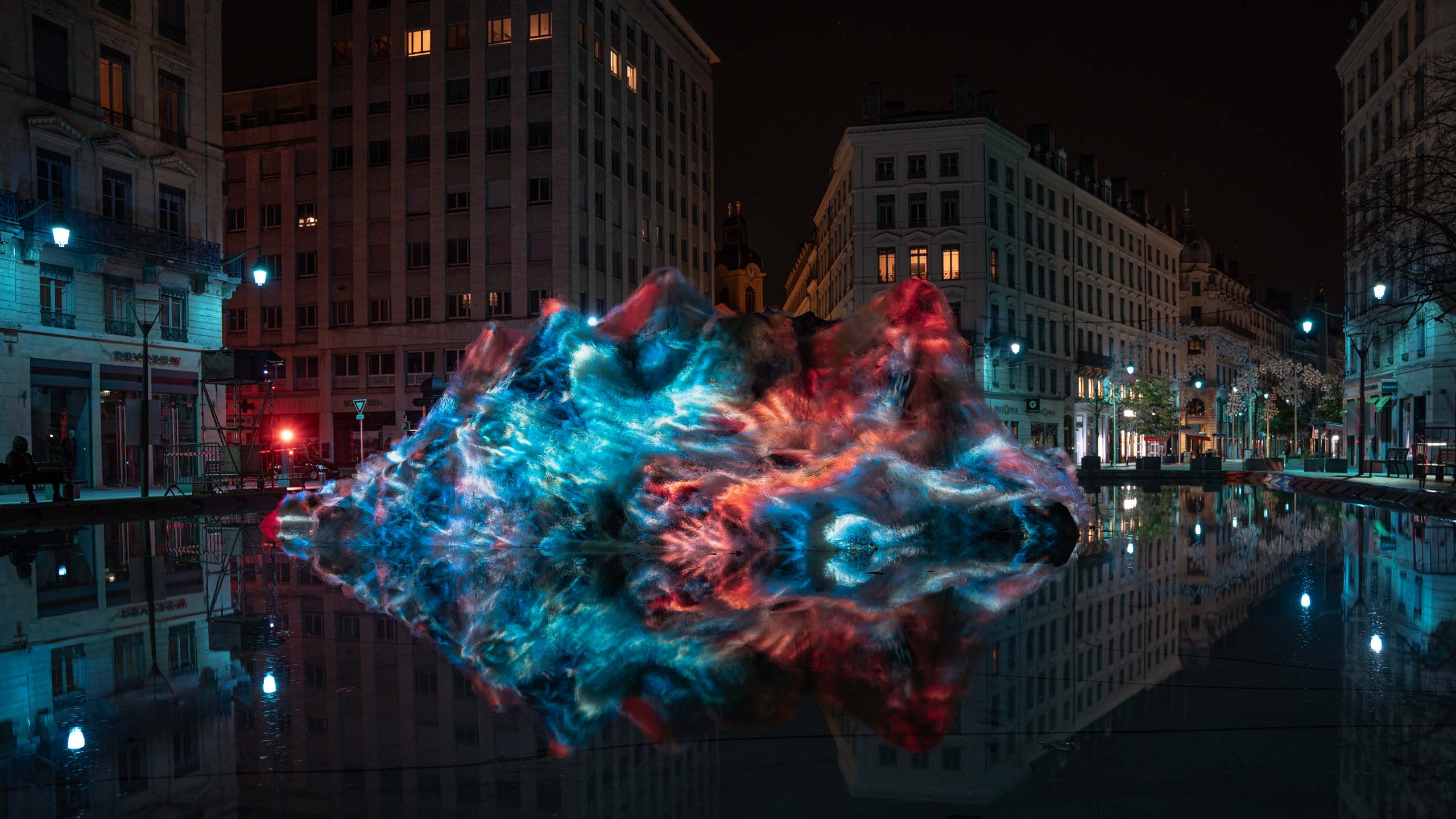 Coral Ghosts Art Installation_Philipp Frank_7 Coral Ghosts light sculpture by Philipp Frank at Fête des Lumières, glowing with deep blue and coral-red projections on ghost nets in urban water setting.